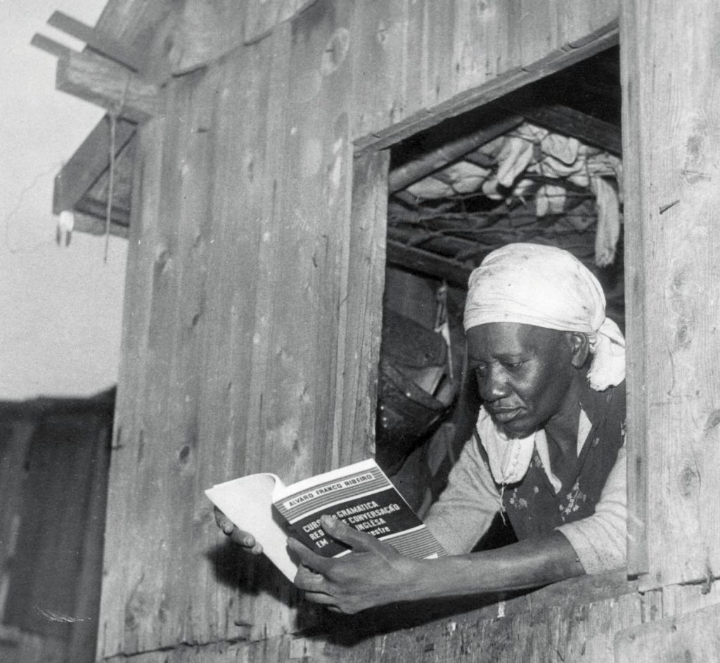 A escritora Carolina Maria de Jesus, mulher negra, veste um pano claro na cabeça e lê um livro na janela de um barraco