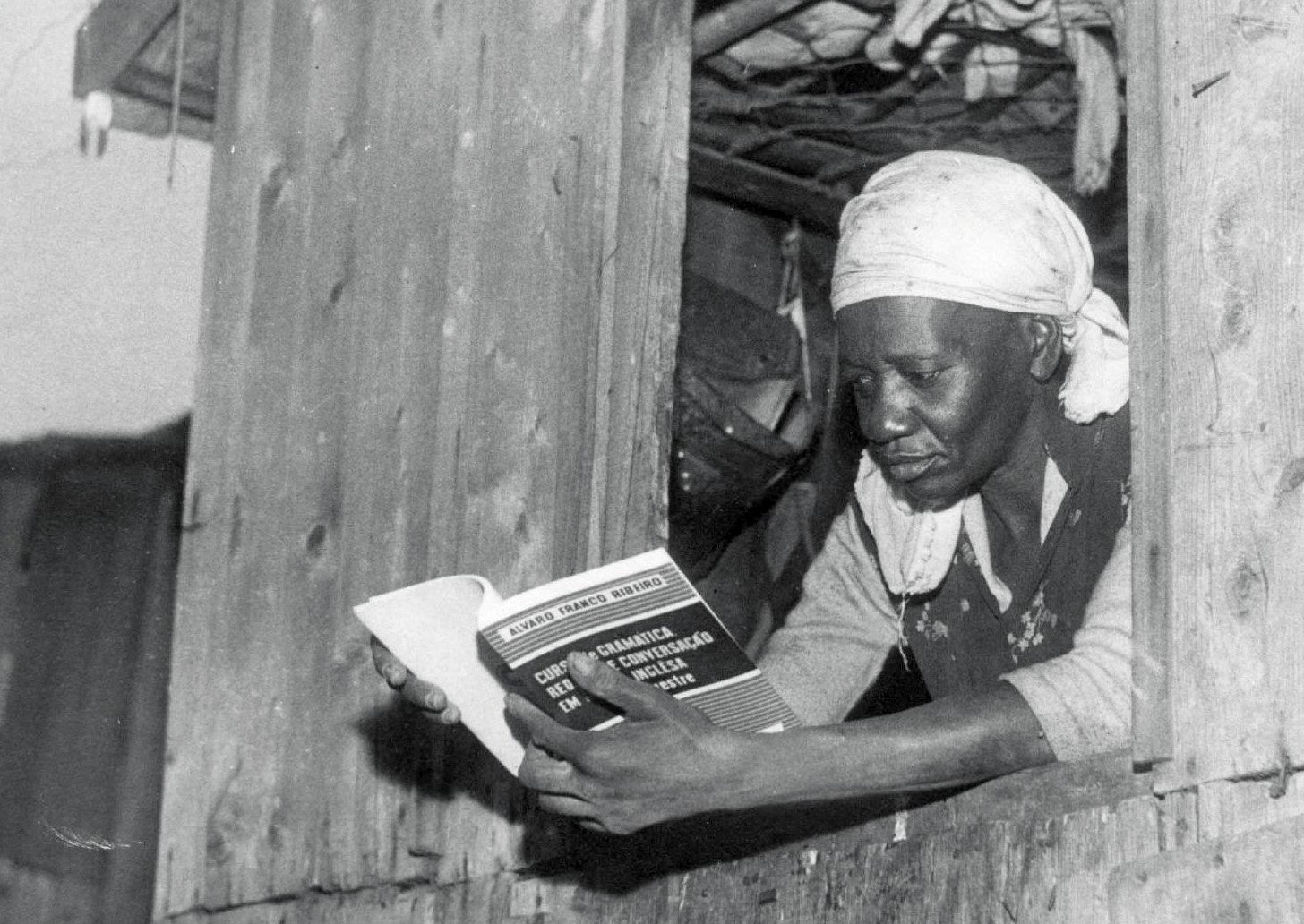 A escritora Carolina Maria de Jesus, mulher negra, veste um pano claro na cabeça e lê um livro na janela de um barraco