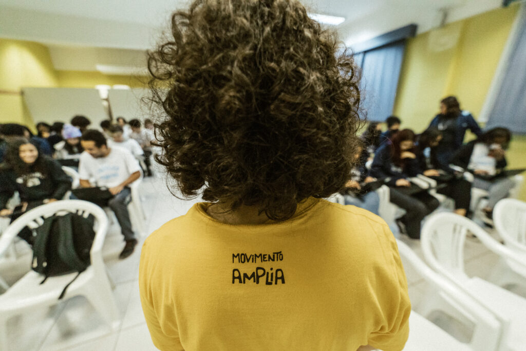 Costas de uma pessoa de cabelos encaracolados que veste uma camiseta amarela com o texto "Movimento Amplia". Ela está de frente pra uma sala de aula